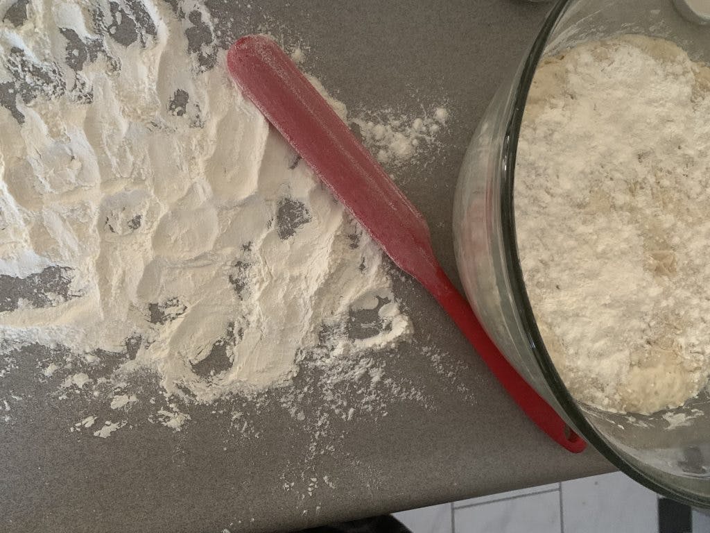 Flour on a work surface with a bowl filled with bread dough and a spatula next to it.